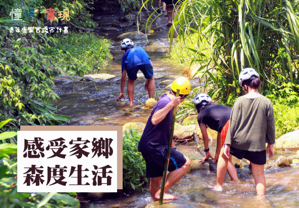 四位參與者在溪流中進行戶外活動，穿著運動服裝和安全帽。左下角標註「感受家鄉森度生活」，右上角為活動單位標誌。畫面展示親近自然、體驗森林生態的主題。