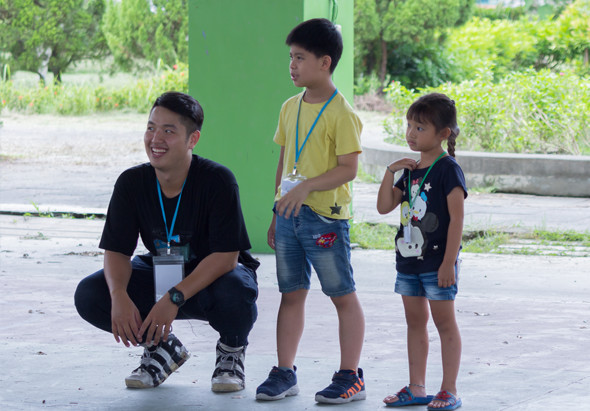 一名配戴識別證的成年男性蹲下與兩名兒童互動，背景為綠色建築物和樹木。三人都面帶笑容，似乎在進行戶外教育或社區活動。