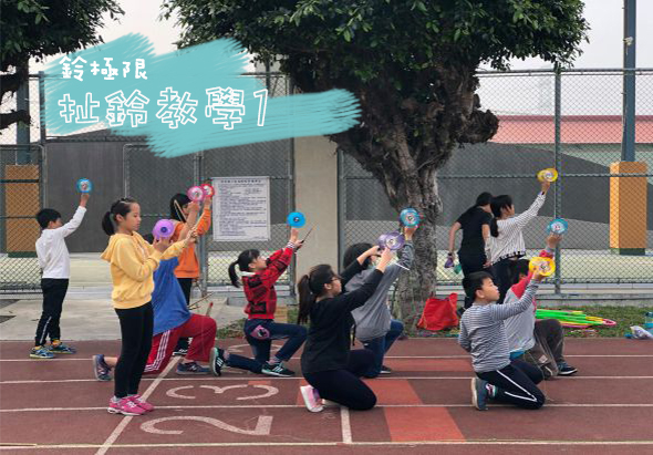學校運動場上，一群小學生穿著彩色運動服，在老師帶領下進行球類運動。孩子們舉起藍色球，做出投擲動作。背景有綠色標語布幔，大樹和運動場設施。場景充滿活力與童趣。