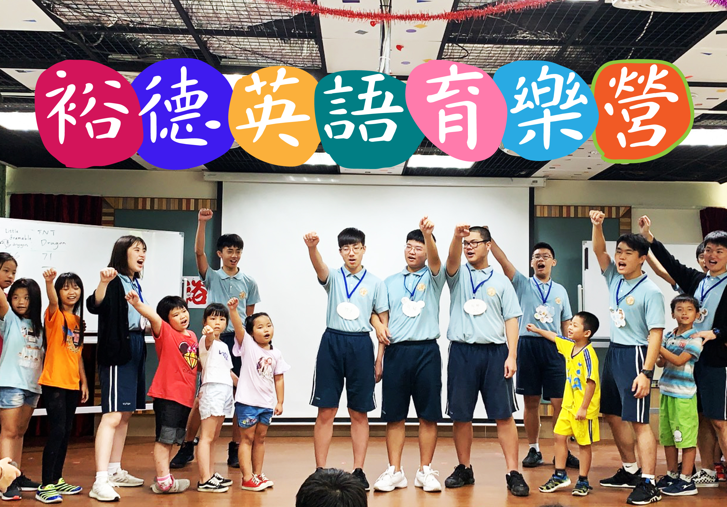 學校禮堂內，一群穿著藍白制服的學生與年幼小朋友開心合唱，舞台上方懸掛彩色圓形標牌，書寫「給德英語首樂營」，現場氣氛歡樂溫馨。