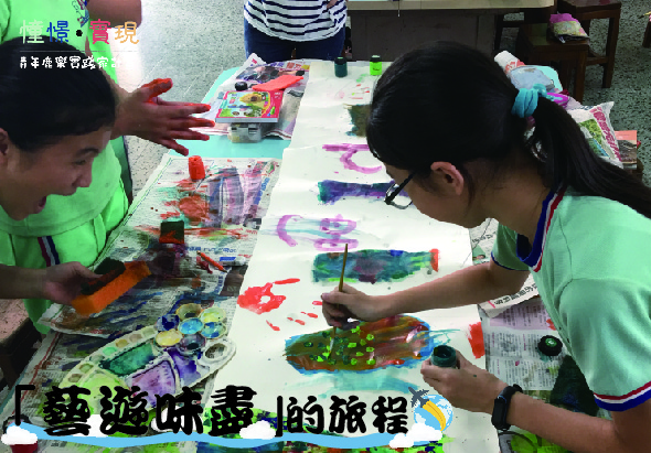 兩名穿著綠色衣服的兒童坐在長桌前進行美術創作，桌上擺放著彩色顏料、畫筆和各種藝術用品，兩人專注地進行繪畫活動。背景可見其他兒童和藝術作品，呈現溫暖的創意教學環境。