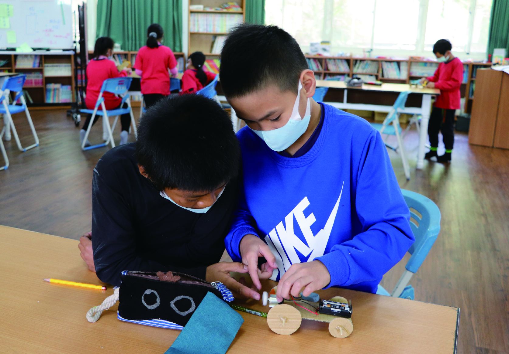 小学生在教室裡進行STEM科學實驗課程，兩名學生戴著口罩，一起操作木製輪子和電子零件組裝簡易機械裝置。背景有其他穿著紅色制服的學生在進行各項活動，室內有書櫃和綠色窗簾。