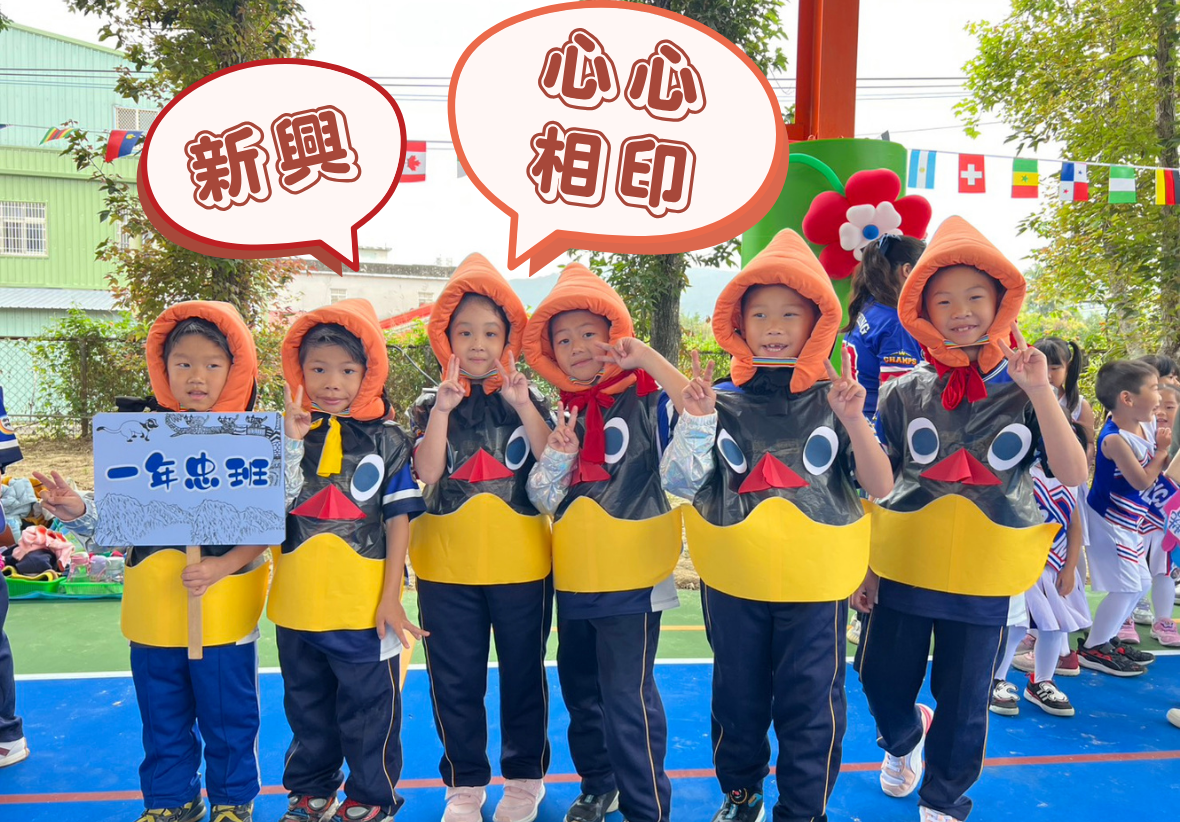 六名小學生穿著黑黃藍三色服裝和橙色帽子，站在學校操場上參加活動。上方有紅色對話框寫著「新興」和「相印」，背景掛著各國國旗。左側學生舉著寫有中文字的牌子，右側可見其他參與活動的小朋友。