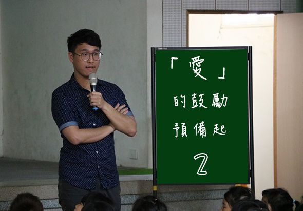 一位戴眼鏡的男性講者手持麥克風在教室進行演講，身後綠色黑板上書寫「愛」的設想與「的設想預備起2」的繁體中文文字，前景可見聽眾的後腦勺。