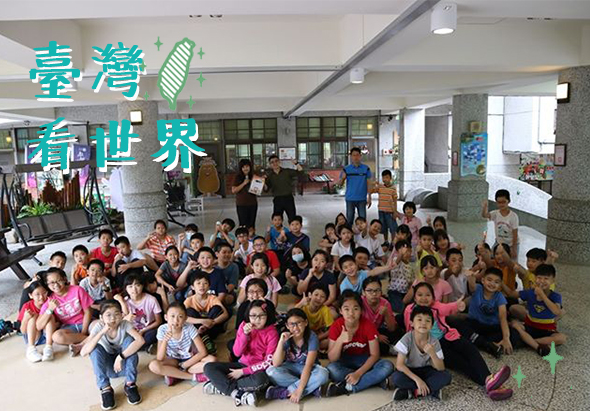 台灣與世界主題活動現場，約百位小學生穿著彩色服裝坐在地上，聚集在室內大廳參與集體活動。背景牆面貼有中英文標題，兩側可見教室門窗和成人工作人員。