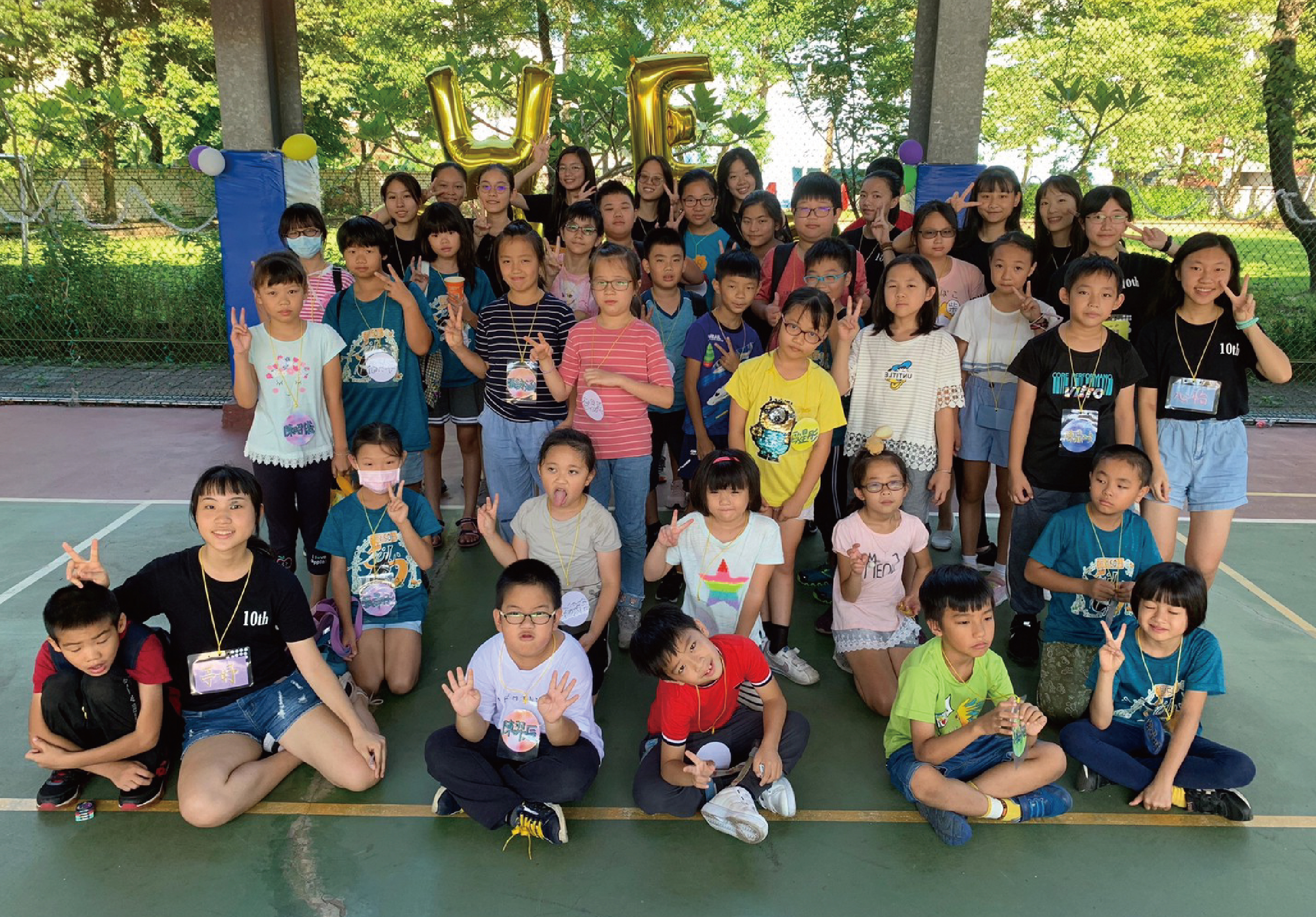 一群小學生在戶外籃球場合照，背景有「UF」金色氣球，樹木和籃球架。學生們穿著彩色衣服，擺出勝利手勢和開心表情，部分學生戴著識別證，呈現歡樂的校園活動氛圍。