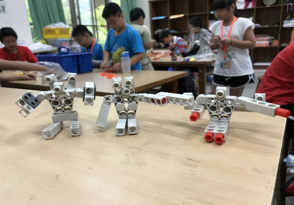 教室內，木桌上放著三個由金屬零件組成的機器人模型。背景中多名學生正在進行STEAM教育活動，操作和組裝機器人零件。窗簾和教室設施可見於背景。