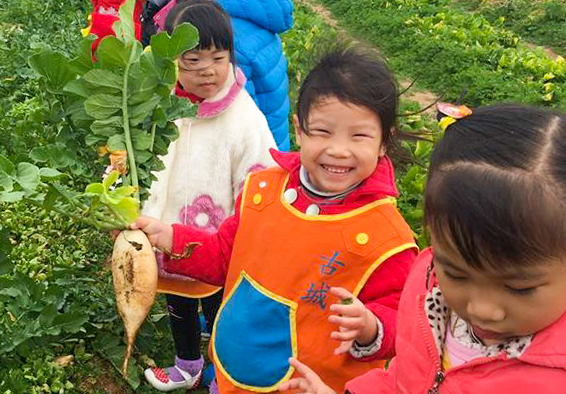 四名兒童在菜園中開心地展示收穫成果，中間女童穿著橙紅色衣服，手持剛挖出的蔬菜。周圍綠意盎然，其他孩童也在觀看和參與農作活動，呈現歡樂的戶外學習場景。
