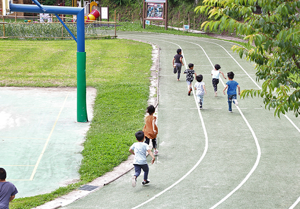 學校操場上，多名小學生正在進行跑步活動。孩子們分散在跑道上奔跑，穿著各色運動服。操場周邊有綠草坪、籃球架和樹木，是進行戶外體育活動的場景。