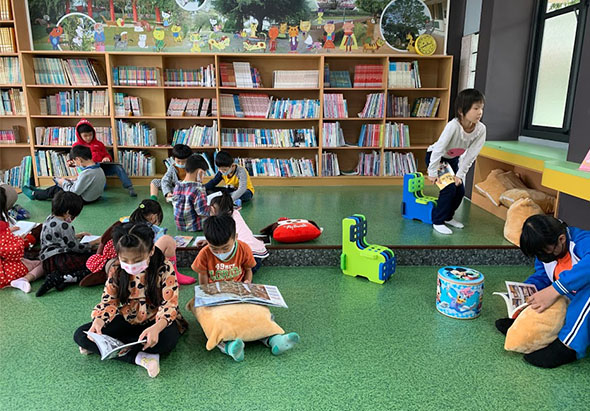 兒童圖書館內，多名幼童坐在綠色地墊上參與閱讀活動。背景是木製書架放滿彩色圖書，教師在旁指導。孩子們手持繪本，專注於故事閱讀，展現溫馨的集體學習氛圍。