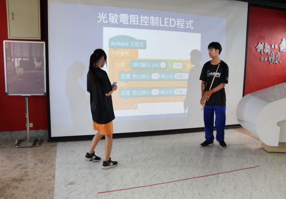 兩名學生在室內進行光敏電阻控制LED程式的展示教學。投影幕上顯示Arduino主控器的電路圖和程式碼框架，包含光敏電阻感測器、LED燈和控制邏輯。背景為教室環境，牆面為紅色。