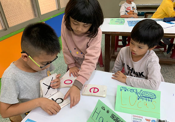 三名小學生在教室裡進行美勞課程，坐在白色桌子前專注地繪畫。左邊男童正在畫ㄱ字母，中間女童和右邊男童各自製作綠色的學習教材，桌上散放著色鉛筆和字卡，背景可見其他學生和教室佈置。
