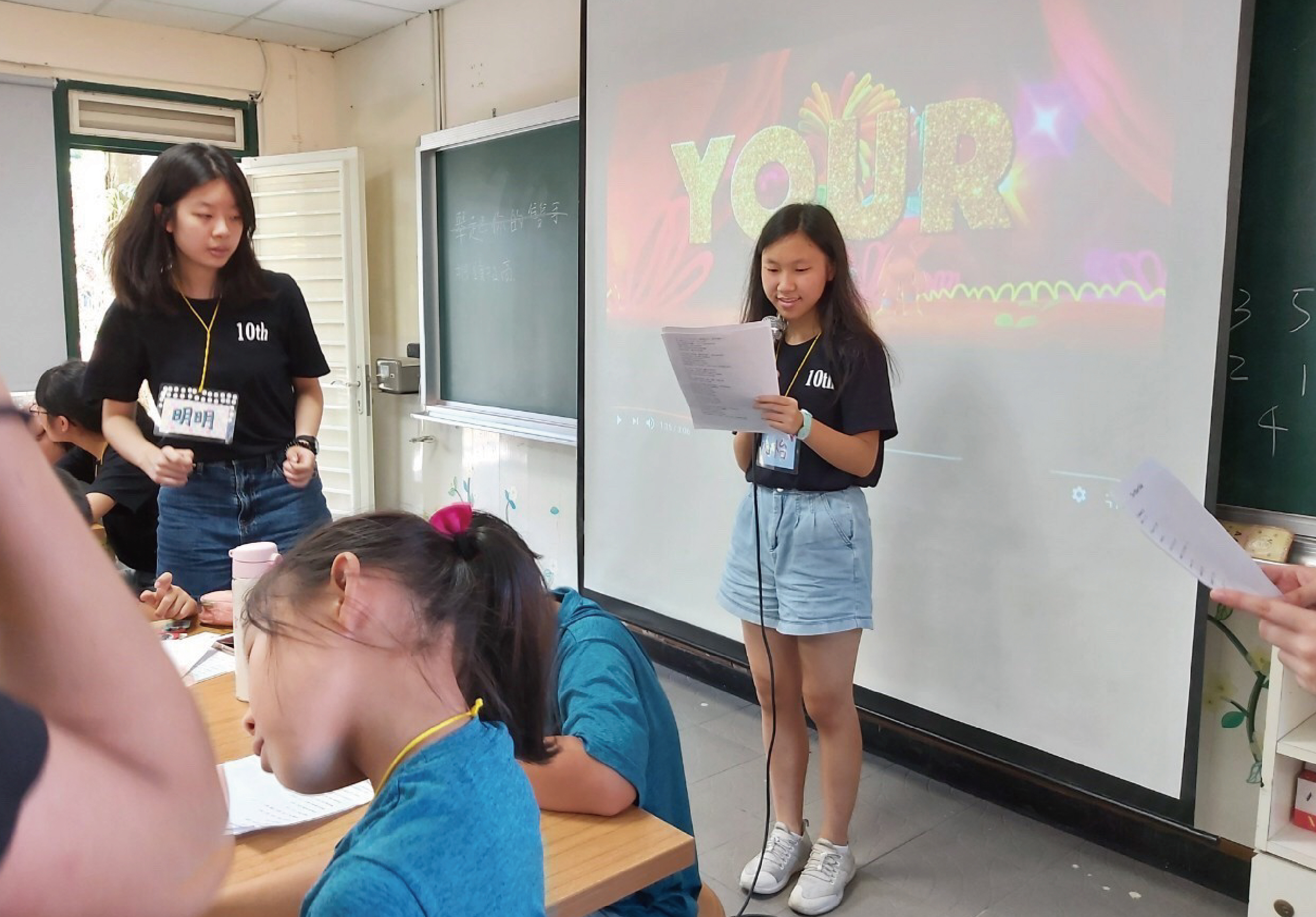 課堂上，兩名穿著黑色10th班服的女學生進行英文簡報。其中一人站在投影幕前，手持講稿，背後投影著彩色的「YOUR」字樣。台下穿著藍色上衣的學生認真聆聽。教室內有黑板、講台和教學設備。