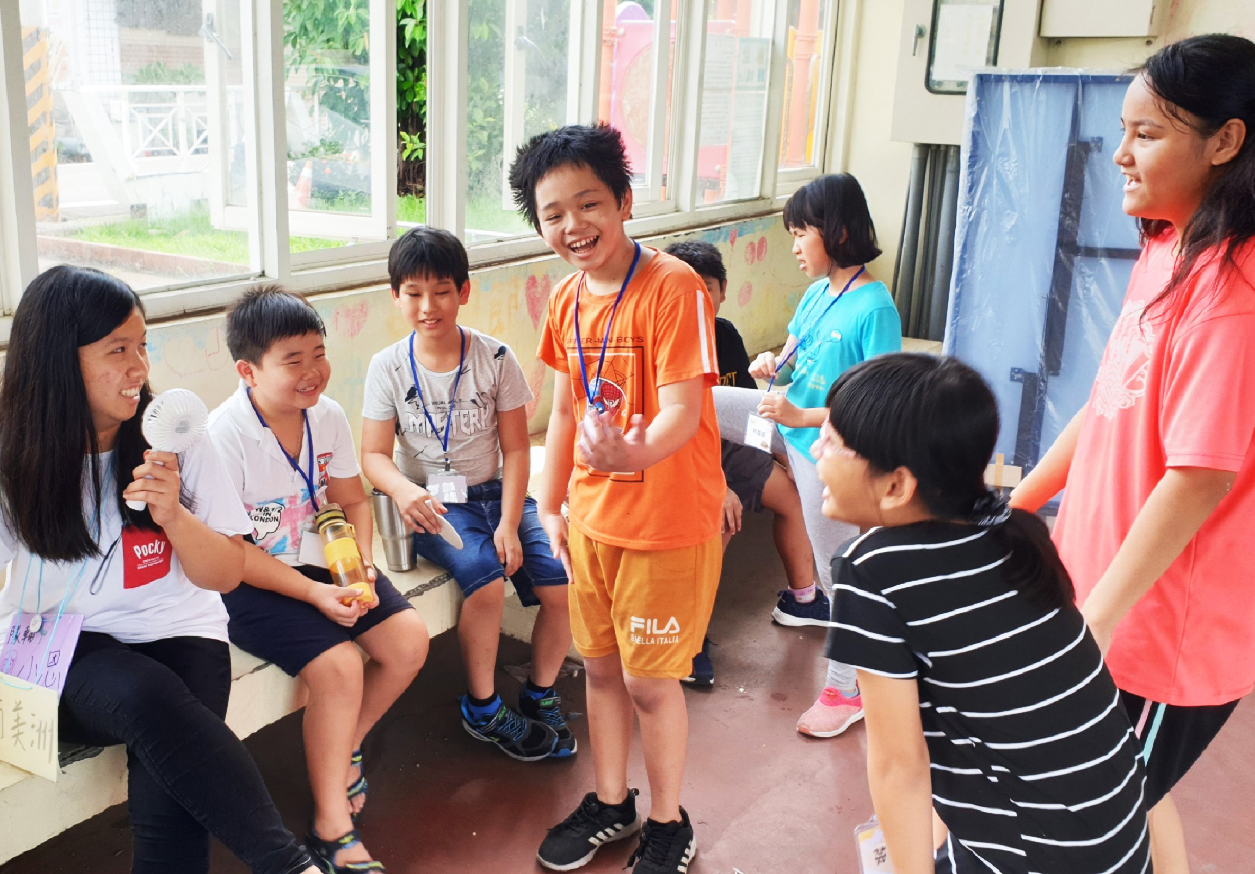 一群小學生在教室內圍成一圈，參與互動遊戲活動。身穿橙色衣服的領隊正在主持遊戲，手持獎品與孩子們互動。孩子們臉上洋溢著歡樂笑容，窗戶外可見校園綠化環境。