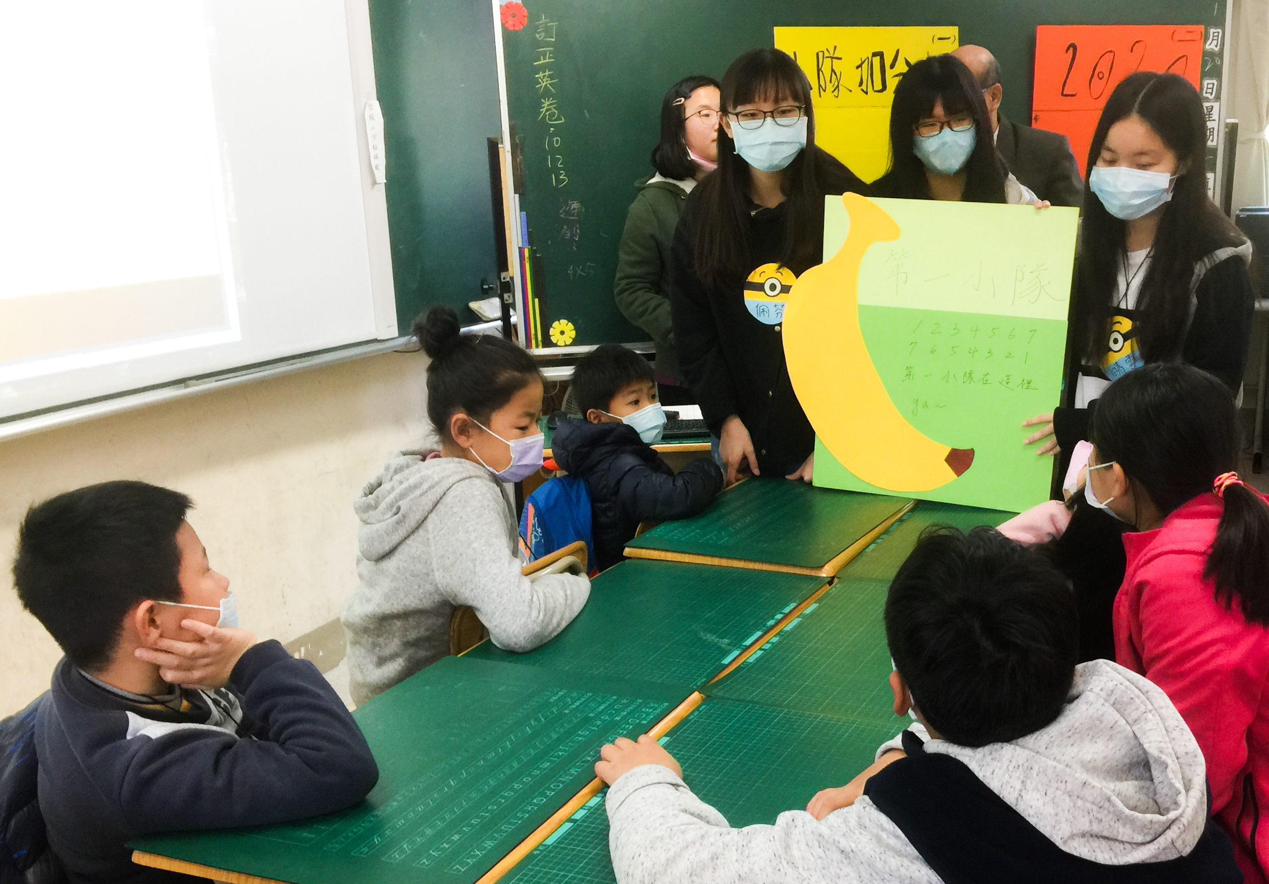 教室內，多位戴著口罩的學生和教師圍坐在綠色課桌旁，共同觀看一張黃綠色的手繪海報。海報上有黃色香蕉圖案和中文文字。黑板上貼著彩色便簽紙，窗邊放有教學用具。