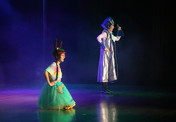 舞台劇表演現場，一位穿著綠色蓬裙、頭戴王冠的年輕演員跪坐舞台左側，面向穿著紫白長袍、頭戴王冠的另一位演員。舞台籠罩在藍紫色燈光中，呈現童話故事場景。