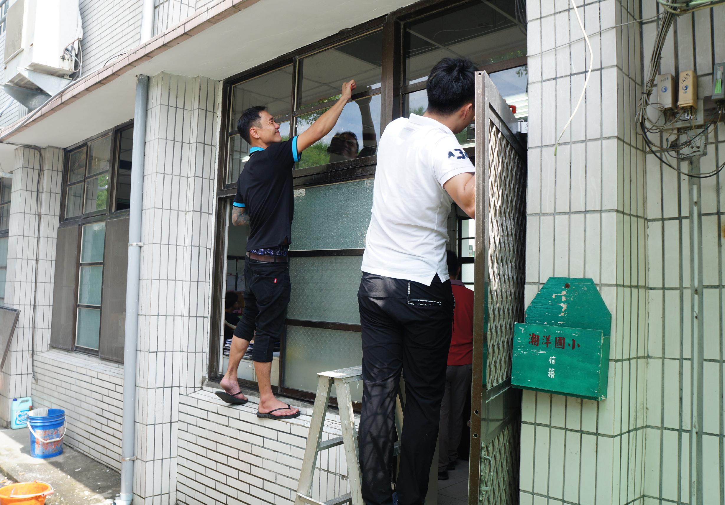 兩名工作人員在建築物外牆安裝或檢修窗戶，其中一人站在梯子上操作窗戶上方，另一人在旁協助。建築物牆面貼有白色瓷磚，右側可見綠色信箱，地面放有水桶和工具。