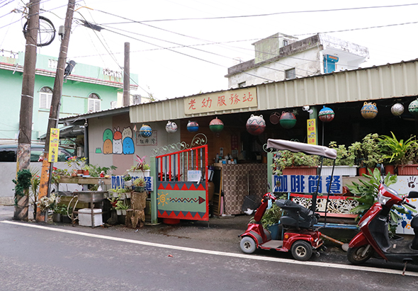 彩色鐵皮屋搭建的幼兒園外觀，門口擺放各式盆栽植物裝飾，牆面繪有卡通圖案。停放著紅色三輪車，背景為老舊公寓建築和電線桿。