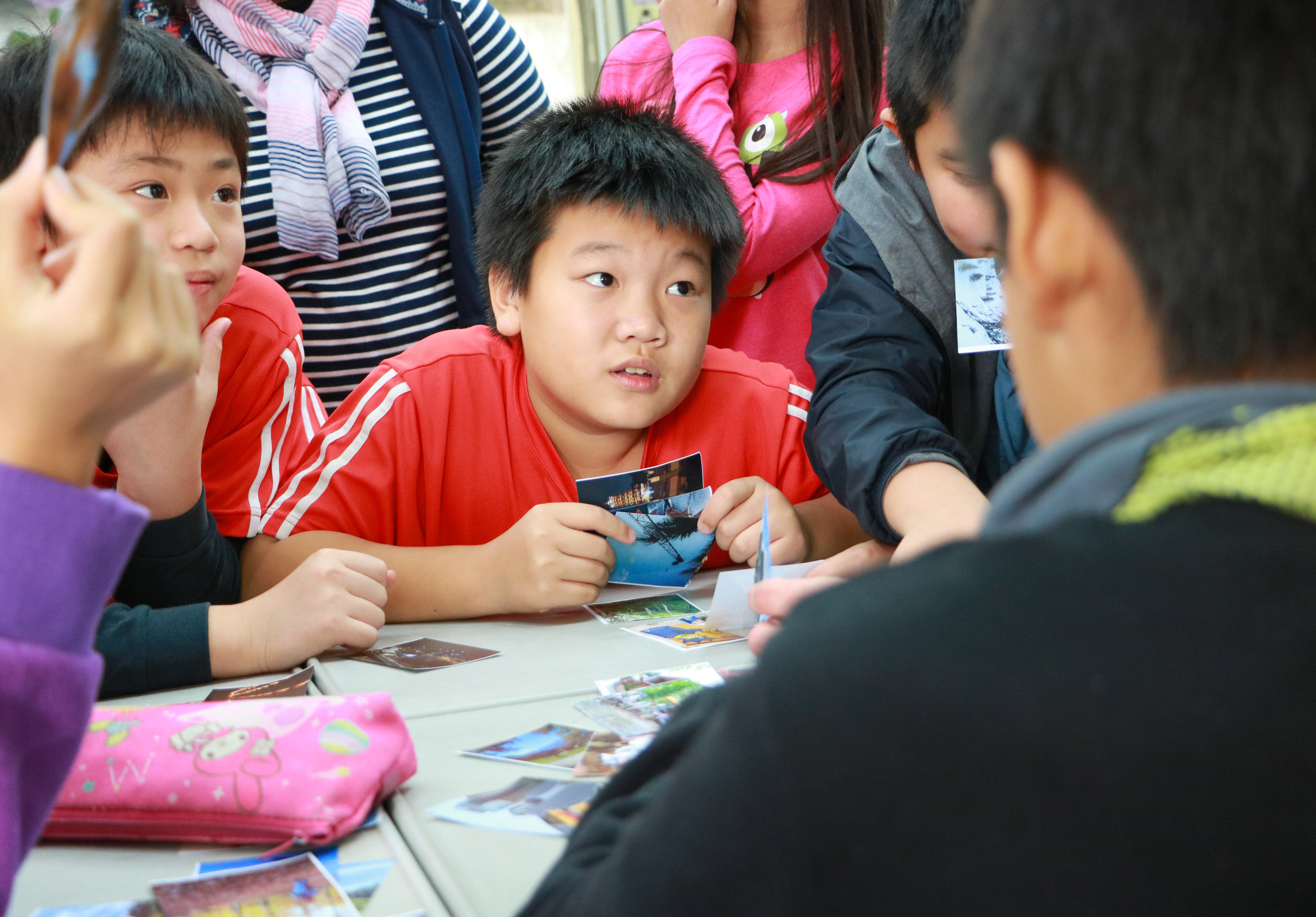 一群小學生圍坐在桌子前，穿著紅色運動服的男童正向其他同學展示手中的明信片或照片。現場有多位家長和老師在旁觀看，呈現溫馨的教室互動場景，小孩們看起來專注投入於分享活動。