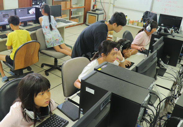 電腦教室內，多名學生坐在電腦桌前進行課程。前景一名女學生專注於螢幕，後方學生在老師指導下進行操作。室內配有多台桌上型電腦、鍵盤滑鼠等設備，背景可見白板和教學用電子設備。