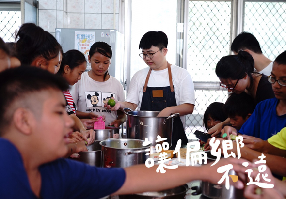 一群年輕人在廚房進行烹飪活動，中央穿著圍裙的講師正向圍在旁邊的參與者示範使用大鍋烹飪。窗戶透進自然光，參與者認真觀看學習。圖片右下角有中文文字水印。
