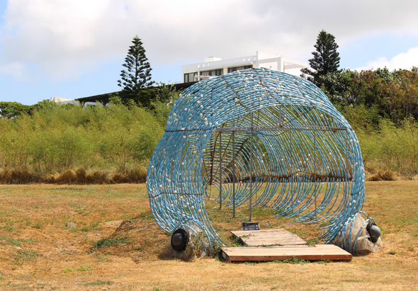 空曠草地上矗立著一個藍色螺旋形金屬裝置藝術品，呈現圓形隧道狀結構，入口處有木製踏板。背景為綠色植被、棕櫚樹和建築物，晴朗天氣下的戶外公共藝術裝置。