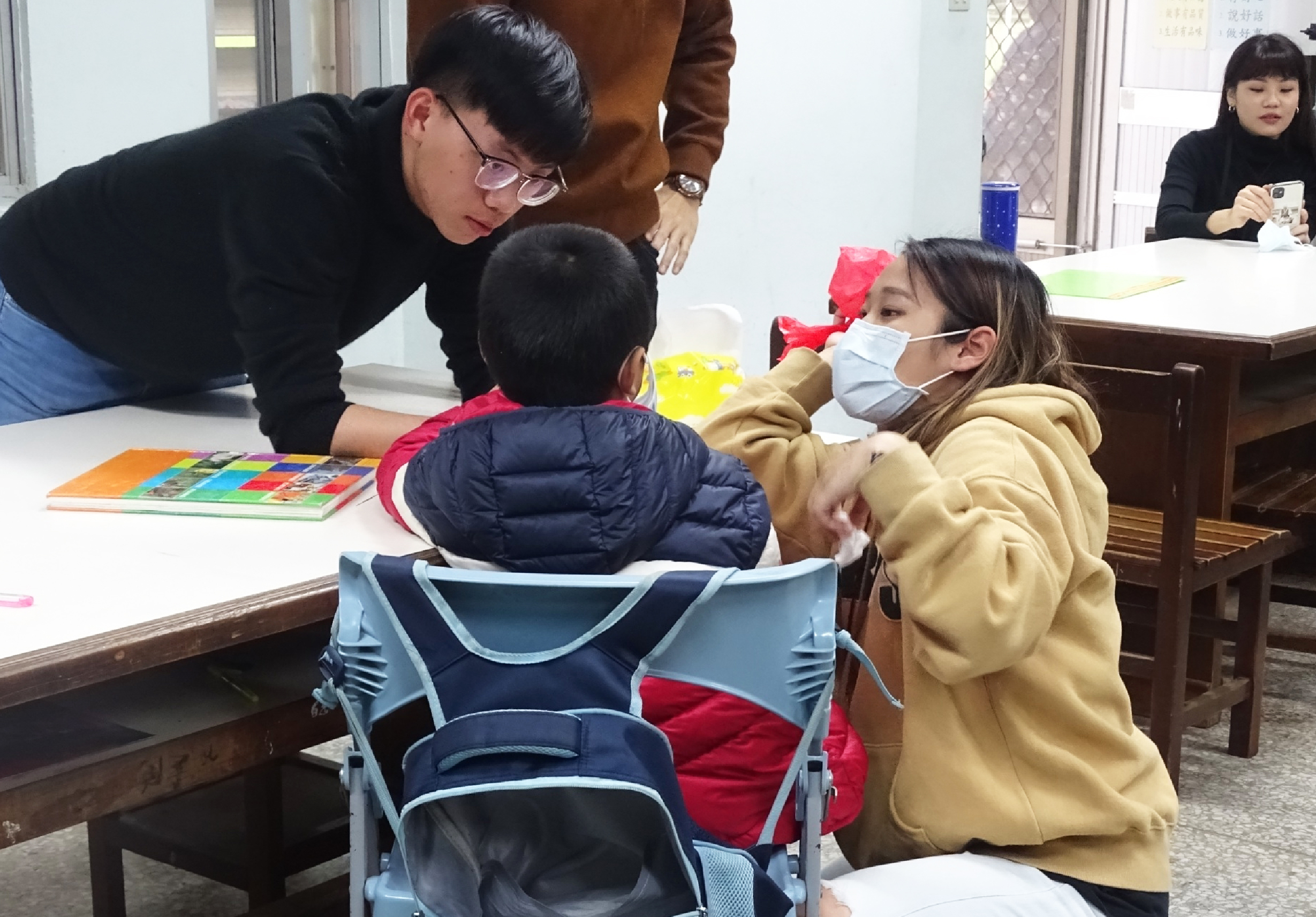 教師在室內教學活動中，彎身指導坐在嬰兒車旁的兩名幼童查看彩色教材書籍，另有兩名女性在旁觀看，桌上擺放多本色彩鮮豔的兒童讀物。