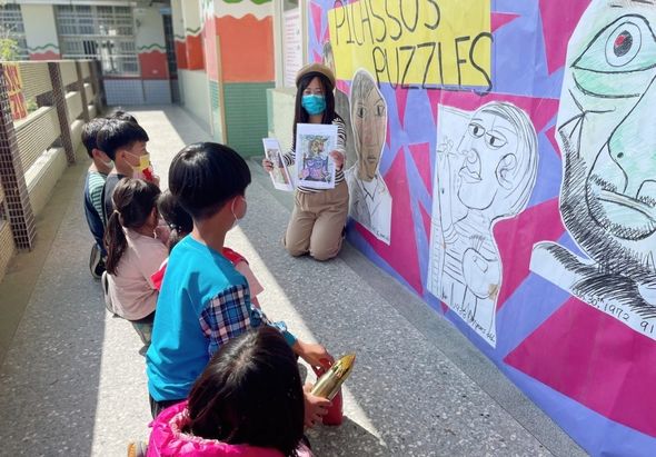 學校走廊內，一位戴著口罩的教師跪坐在地上，向圍坐的五位學童展示與講解彩色壁畫。壁畫上繪有多張卡通人物肖像，右側牆面寫著「FAMOUS PUZZLES」字樣。這是一場藝術教育活動。