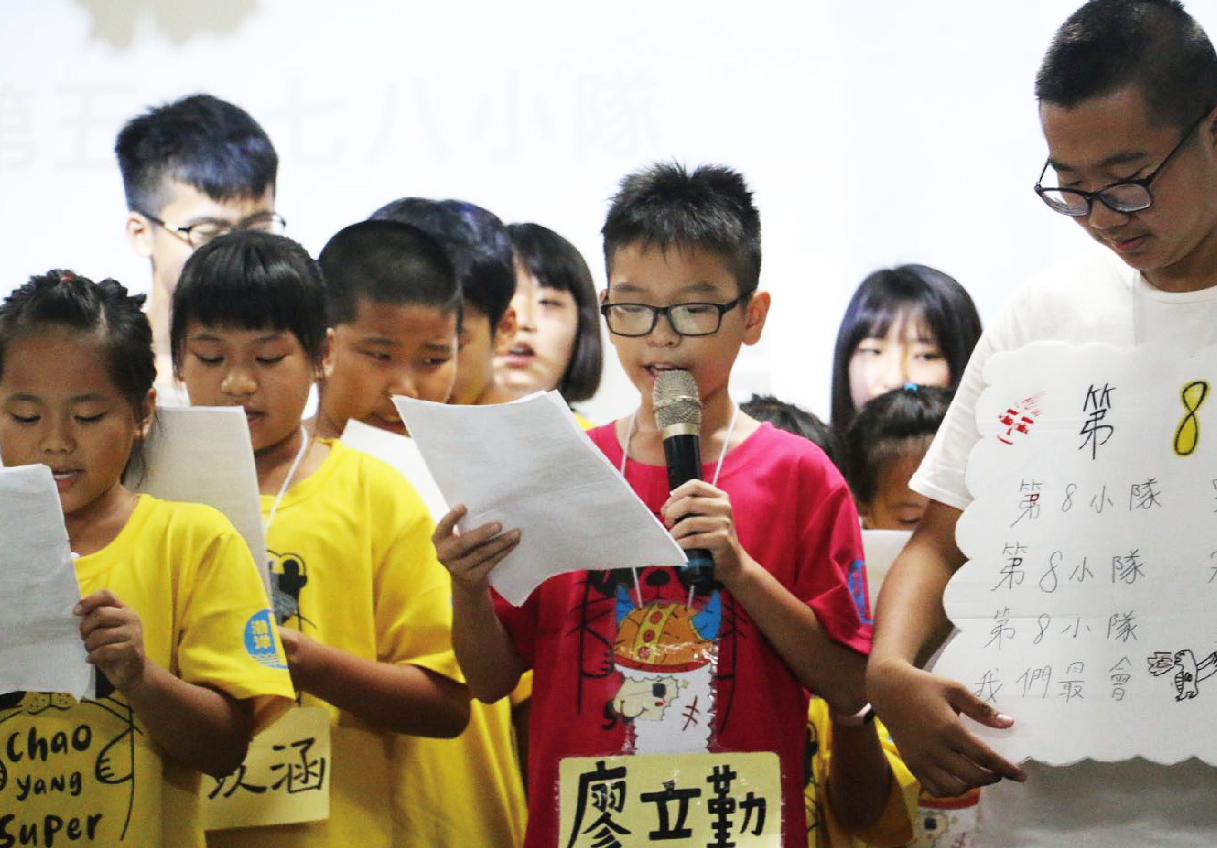 一群小學生穿著黃紅色制服，手持紙張進行表演或朗誦活動。中央穿著紅色衣服的男孩拿著麥克風，身旁的成人協助舉著寫有中文字的白色紙張，背景為白色牆面。