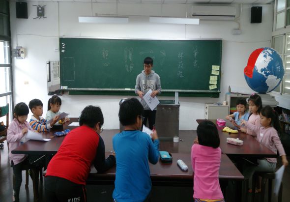 小學教室內，一位教師站在黑板前向學生授課，黑板上寫有中文字。約十名學生穿著粉紅色和彩色衣服坐在課桌前聽講。教室右側牆上掛著彩色地球儀。窗戶採光明亮，牆上有揚聲器和教學用具。