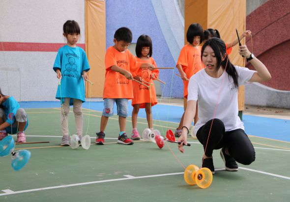 室內運動場地上，一位女性教師蹲身示範悠悠球技巧，身穿白色上衣和黑色褲子。周圍穿著藍色和橙色運動服的兒童聚集觀看學習，現場擺放多個悠悠球，背景為彩色牆面和運動設施。