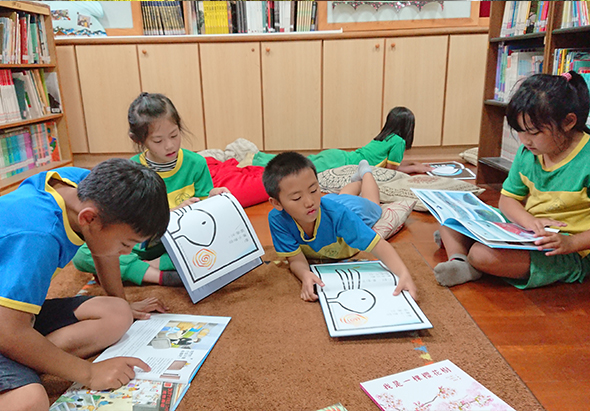 圖書館內，穿著綠黃色和藍色制服的小學生們坐在地板上，圍成一圈進行閱讀活動。他們正在閱讀繪本和著色畫冊，專注地看著手中的書籍。背景是木製書架，擺放著各式兒童圖書。
