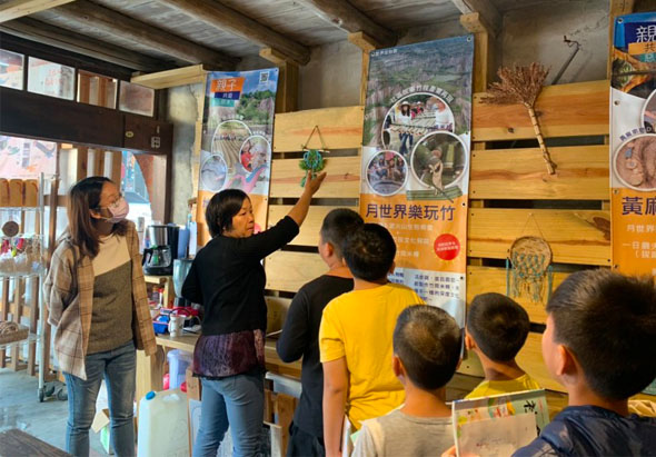 室內教育展示空間，一位女性講師向6名兒童介紹木製展示板上的旅遊海報和景點照片，牆上張貼著繁體中文旅遊宣傳資訊，呈現互動式的地方文化推廣活動。