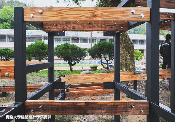 校園戶外木製涼亭結構，由黑色支柱與木質橫樑組成，內部配有木製座椅。背景可見綠化校園、樹木及建築物。一名穿著黑色上衣的人物站在涼亭右側。