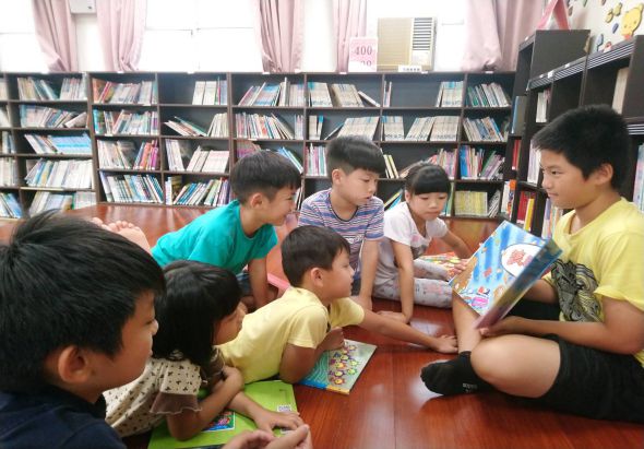 圖書館內，一位穿著黃色衣服的年長志工坐在地板上，與七名小學生圍坐成圈，一起閱讀和分享圖畫書。背景是擺滿書籍的書架和粉紅色窗簾，呈現溫馨的兒童閱讀推廣場景。