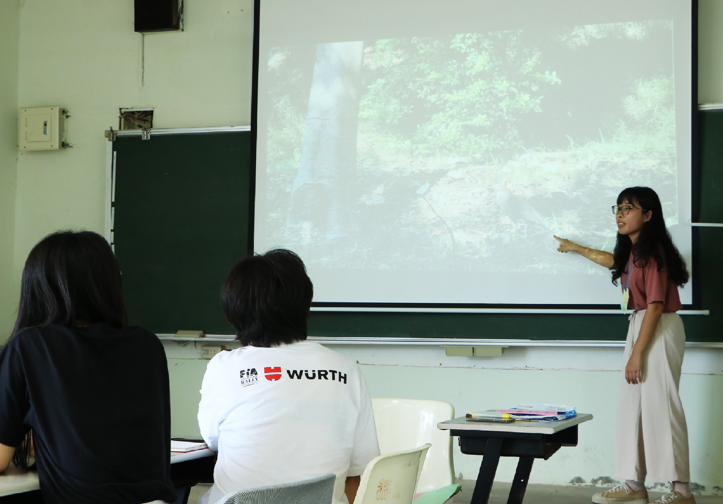 課堂教學場景，一位女學生站在講台前，穿著印有「WÜRTH」標誌的白色T恤，指向投影螢幕上的森林影像進行解說。兩位同學坐在教室前排聆聽，講台上擺放著教科書和文具用品。