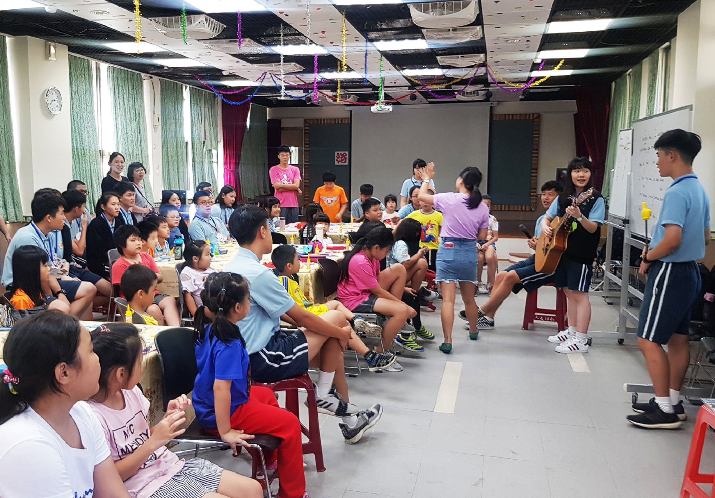 一場室內音樂教學活動，數十位小學生坐在椅子上圍成半圓形，一位女性音樂老師手持吉他站在中央演奏教學。教室天花板裝飾著彩色的紙條和飾品，牆面布置有綠色窗簾和紅色布幔。參與者認真聆聽和觀看表演。