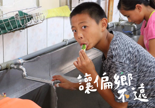 一名穿著灰色短袖上衣的男童在廚房水槽前刷牙，身後有其他學童。廚房牆面貼有綠色磁磚，水槽上方掛著黃色毛巾和瀝水籃。圖片右上角有白色文字「港僑鄉不選」。