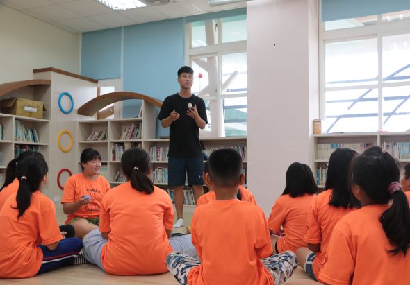 一位男性講師站在圖書館中央，面向坐成一圈穿著橙色制服的學童進行教學。背景有書架、大窗戶和藍色牆面，現場進行著互動式的教育活動。