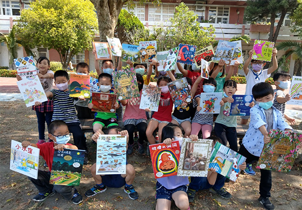 一群小學生在校園空地舉行閱讀分享活動，眾多兒童舉起各式彩色繪本和書籍展示，背景為校舍建築和綠樹，現場充滿歡樂的閱讀推廣氛圍。