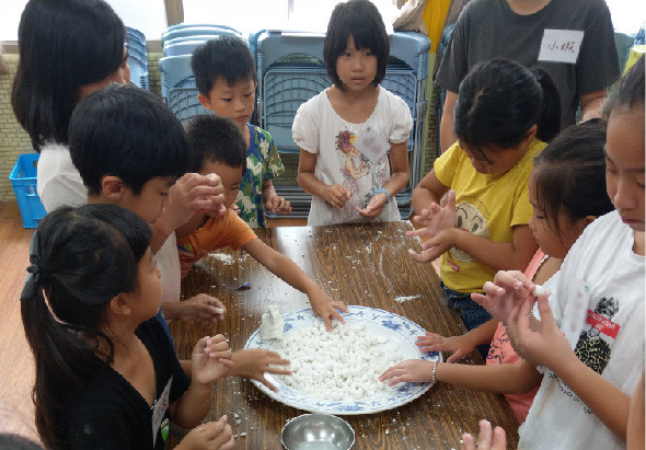 一群小學生圍著木製餐桌，用雙手在白色盤子裡揉搓米粒，進行食物製作或感官體驗活動。室外場景，背景可見輪胎和教室環境，展現互動式學習的教育活動。