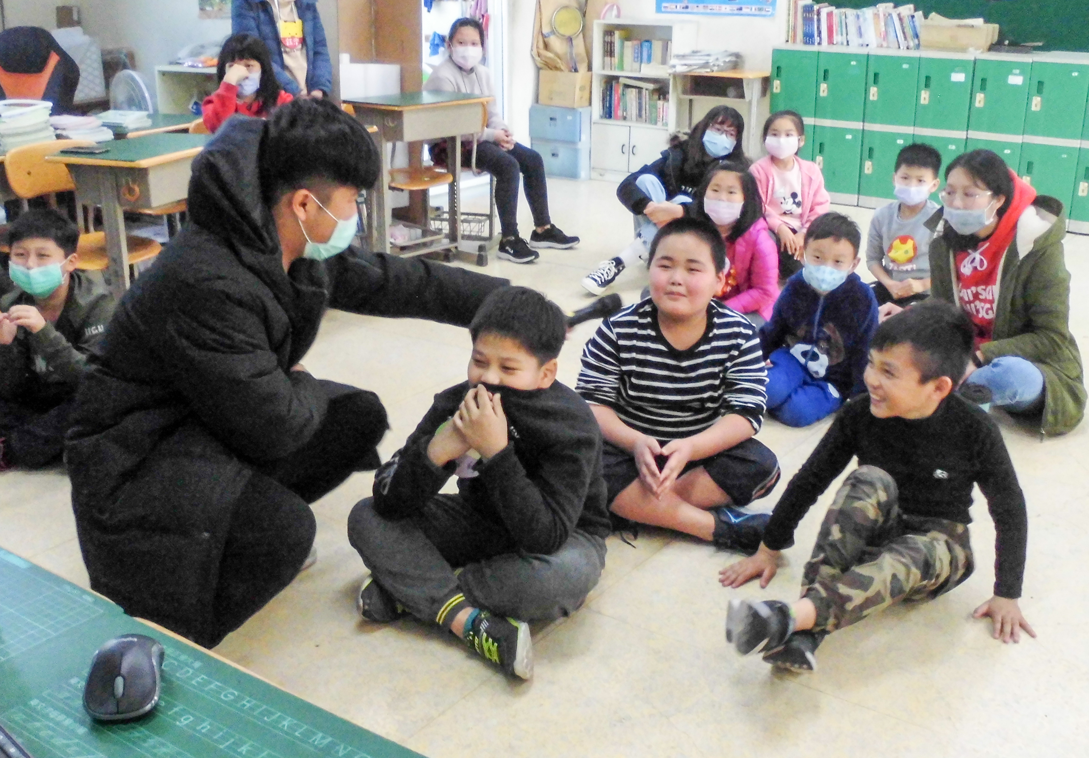 教室內一位年長學生蹲坐在地上與多名年幼學生進行互動教學或遊戲，學生們都戴著口罩，坐成一圈圍繞著講者，背景可見書架、綠色置物櫃和教室環境。