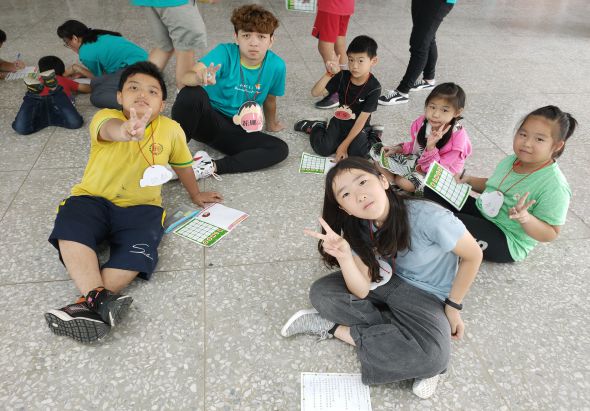 一群小學生坐在室內地板上，手持學習單或教材，圍成一圈參與課堂活動。孩子們穿著各色衣服，認真地看向鏡頭或低頭查看手中的資料，背景可見站立的大人和其他學生。
