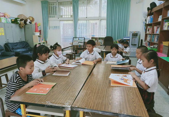 小學教室內，約十名穿著白色制服的學生坐在木製長桌前閱讀書籍。教室配有書架、綠色窗簾、講台和教學用具，學生認真專注於閱讀活動。