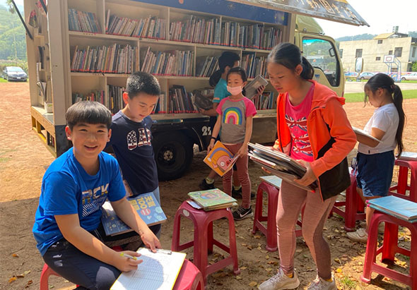 一輛流動圖書館停靠在戶外，幾名兒童坐在紅色小凳子上，向一位女性志工借閱書籍。兒童手持圖書，展現閱讀興趣。背景可見書籍滿載的貨車書架，以及建築物。這是一場鄉村地區的閱讀推廣活動。