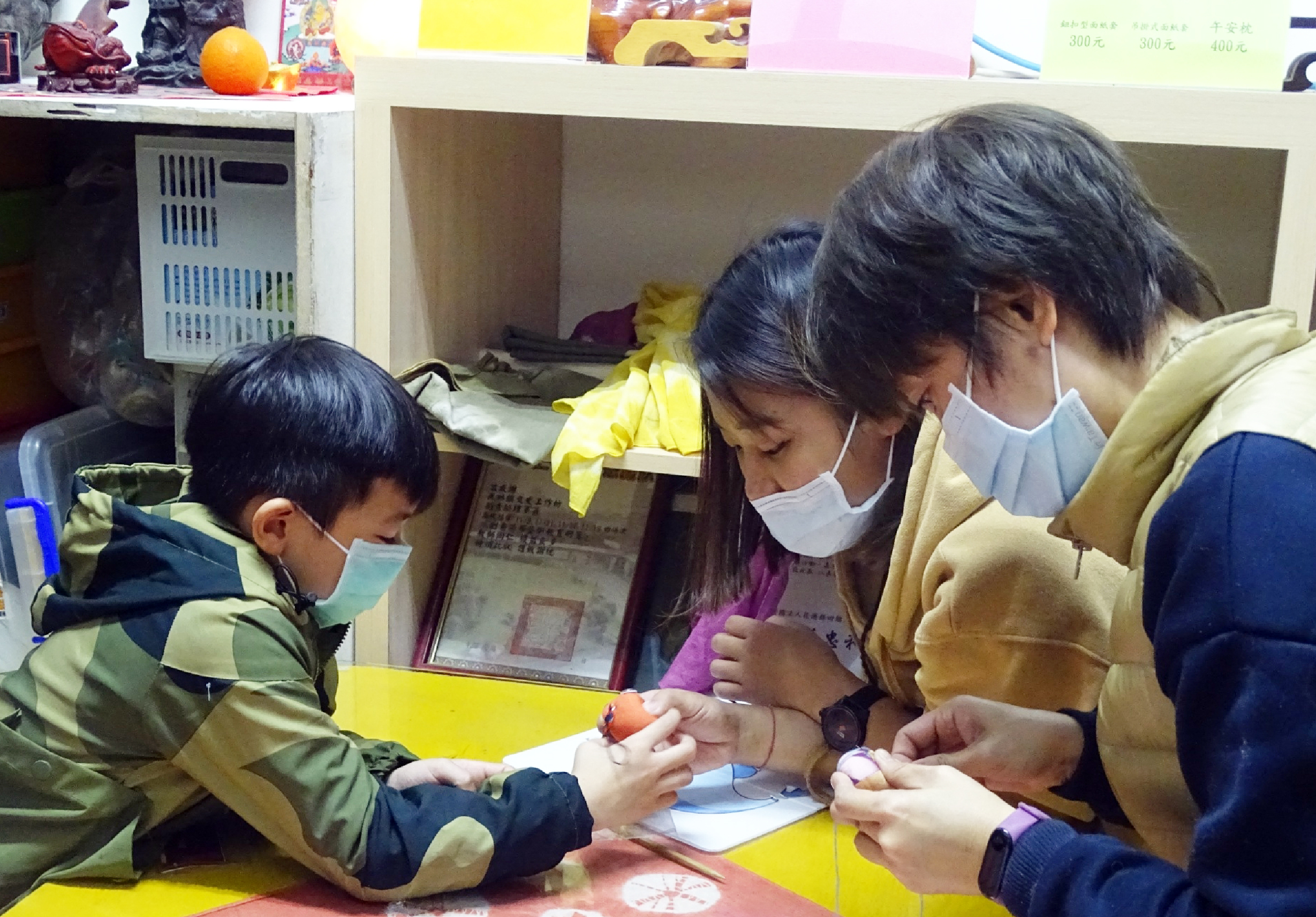 室內教室場景，一位戴口罩的女性教師與三位戴口罩的小學生圍坐在黃色桌邊，正進行藝術創作或手工活動。學生專注地在紙張上作畫或書寫，教師在旁指導。背景可見櫃子、教室佈置和教材。