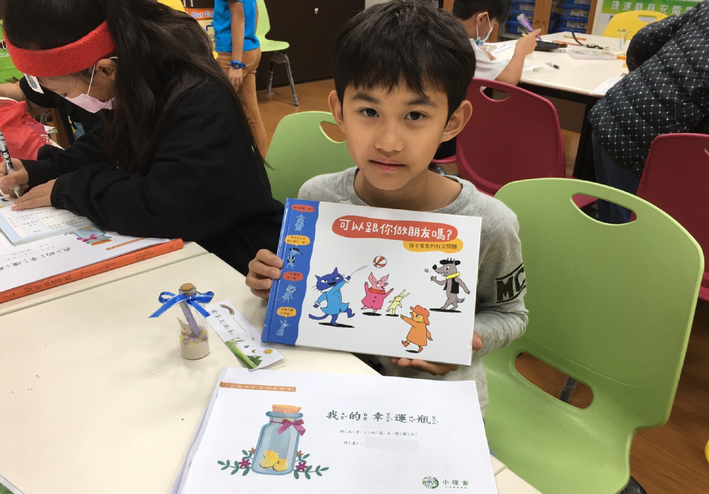 小男孩在教室裡展示一本兒童圖書，書籍標題為「可以超越你做朋友嗎？」，封面繪有卡通角色。桌上擺放著練習作業和文具，身後有成人和其他學童進行閱讀活動。