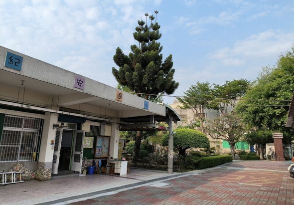 街道上一座現代化車站建築，白色外牆配有多個標誌，前方有遮陽棚。站前廣場鋪設紅磚地面，種植修剪整齊的綠色植栽和高聳的柏樹，周圍環境整潔優美。
