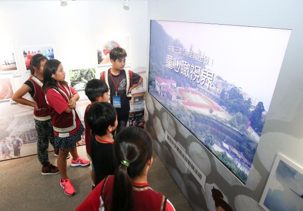一群穿著紅色背心的小學生在展覽現場觀看大型螢幕展示。螢幕上播放著山區建築群的風景影像及中文字幕介紹。講解員站在旁邊為學生進行導覽解說。