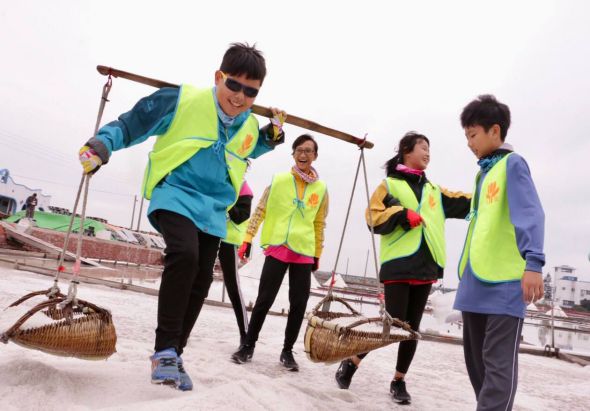 五名穿著螢光黃背心的兒童在濱河地帶進行環保活動，他們扛著竹製農具和籃子，模擬傳統漁業勞作，展示環境保護和文化體驗的教育活動。